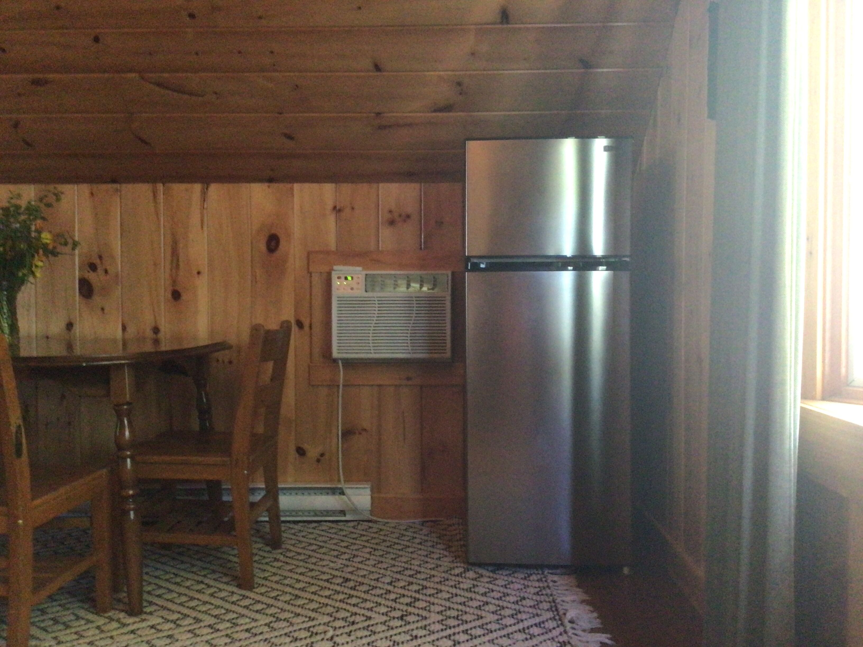 A compact kitchen with wooden walls, a stainless steel refrigerator, a wooden dining table with chairs, and a patterned rug.