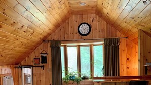 A spacious room with wooden walls and a sloped ceiling, featuring a large window with curtains, a wooden bar area, and a clock on the wall. - Down the road from Bayview Beach on Ferry Rd! Bright, and newly furnished 2 BR. (Saco)