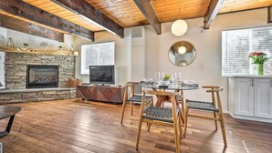 A spacious living room with a stone fireplace, wooden ceiling beams, a round mirror, and a dining area with a glass table and wicker chairs.