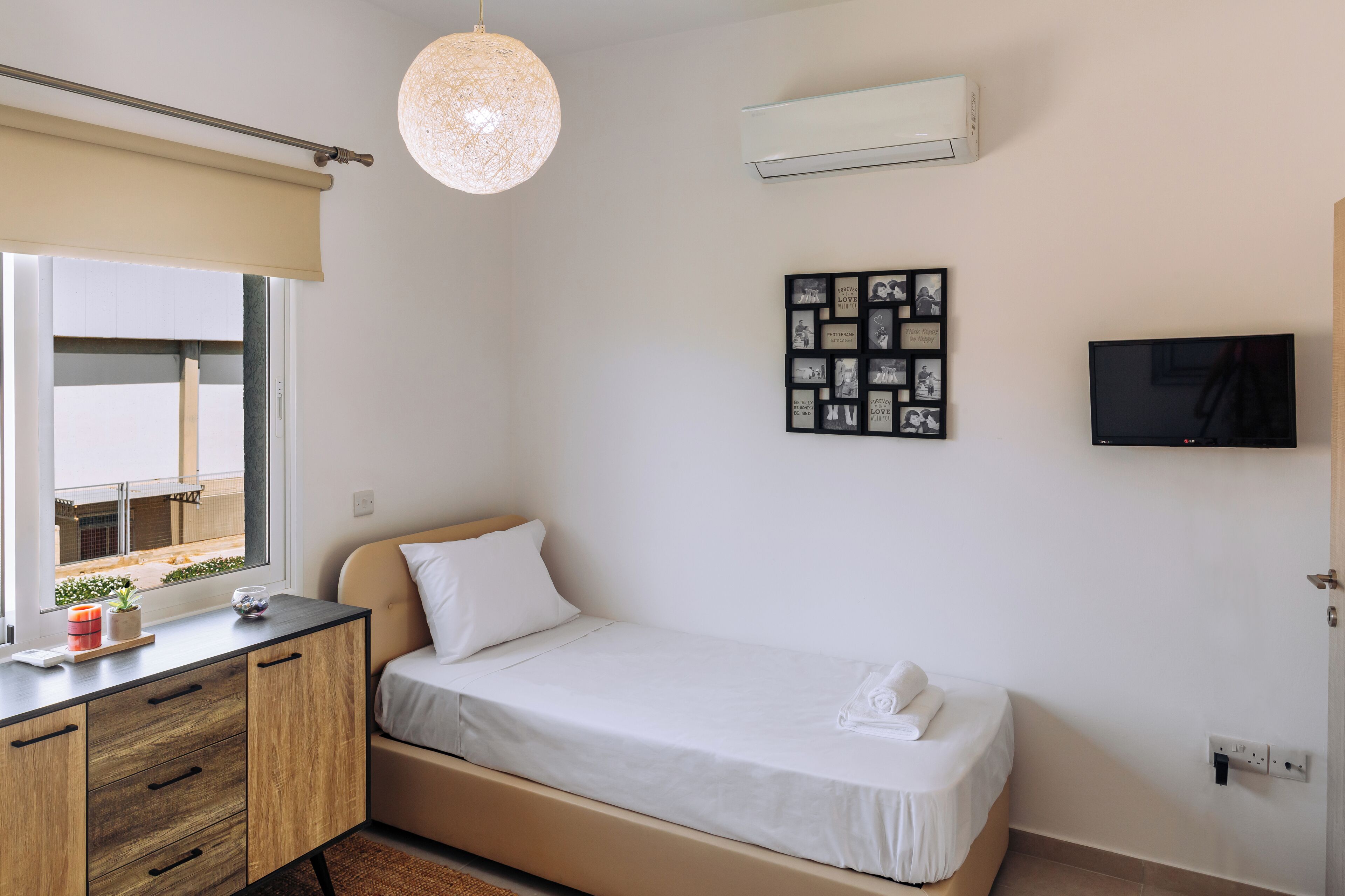 A bedroom with a bed, a desk, a hanging light fixture, a wall-mounted air conditioner, and a framed photo collage.