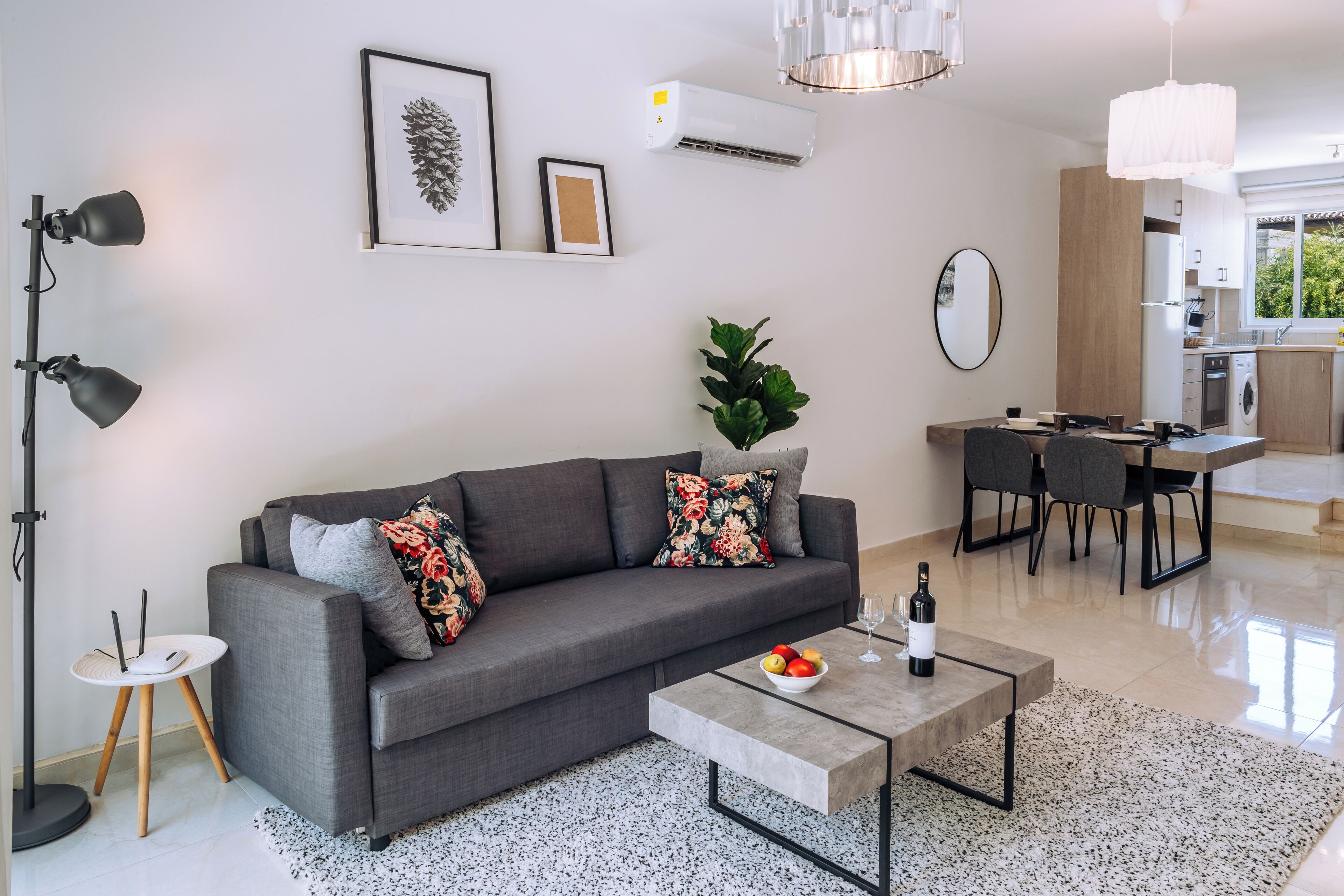 Una sala moderna con un sofá gris, una mesa de centro con una botella y vasos, un comedor con una mesa y sillas, y una cocina al fondo.