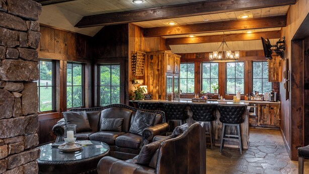 L’intérieur d’un chalet rustique, avec un mur en pierre, des meubles en cuir, un coin bar et de grandes fenêtres.