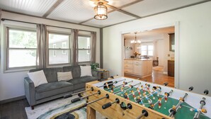 En stue med et bordtennisspil, en grå sofa og et køkken i baggrunden.