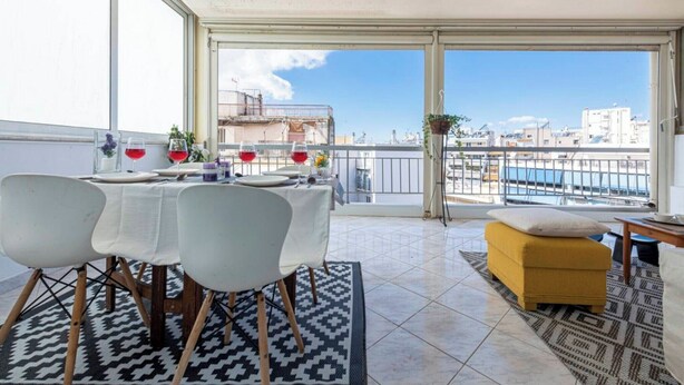 A balcony with a dining table set for four, a yellow ottoman, and a view of the cityscape. - "cityscape-zen-penthouse" (Vyronas)