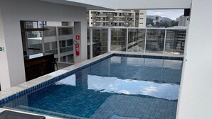 Piscina no terraço com azulejos azuis translúcidos, duas mesas de vidro e um espaço de bar com banquetas.