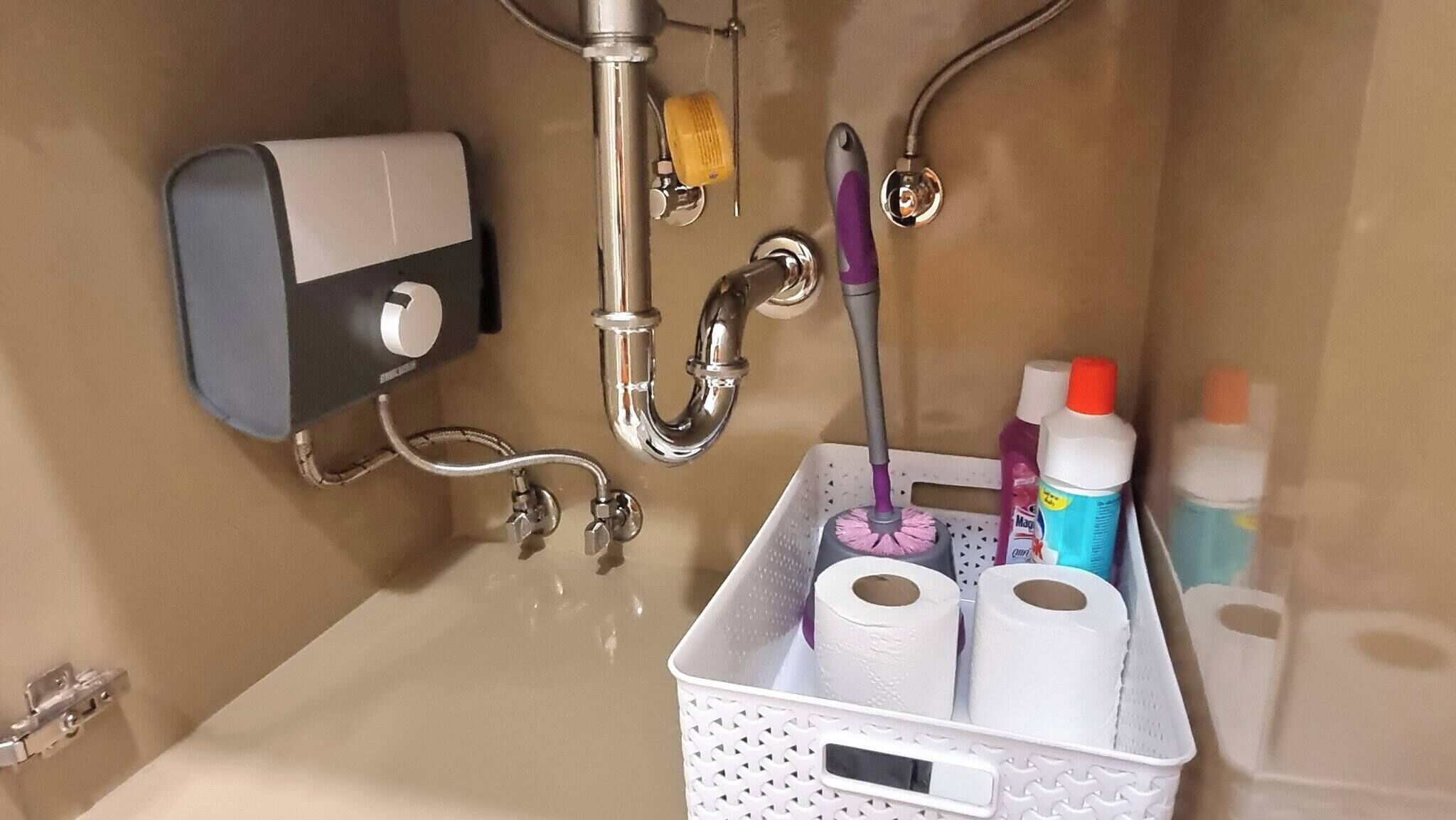 Un lavabo de salle de bain équipé d’un distributeur de savon, d’un robinet, d’une brosse à main et d’un panier contenant du papier toilette et des produits de nettoyage.