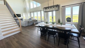 En rummelig opholdsstue med en trætrappe, et spisebord med stole og store vinduer med udsigt til omgivelserne.