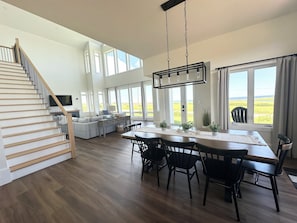 A spacious living area with a wooden staircase, a dining table with chairs, and large windows offering a view of the outdoors. - 4 Bedroom Luxury Ocean View Beach House, Minutes To North Shore Beaches (Park Corner)