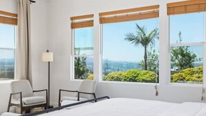 Habitación de hotel con cama, dos sillas, una lámpara de pie y ventanas amplias que ofrecen vistas a zonas verdes y un paisaje urbano.