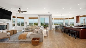 Un salón amplio con chimenea, asientos cómodos, una cocina con barra y ventanas grandes que ofrecen vistas.