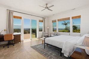 A bedroom with a large bed, a desk, and a chair, with a view of the outdoors through large windows.