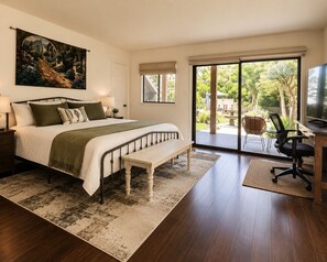 A bedroom with a large bed, a bench, a desk with a chair, a TV, and a view of a garden through a sliding door.