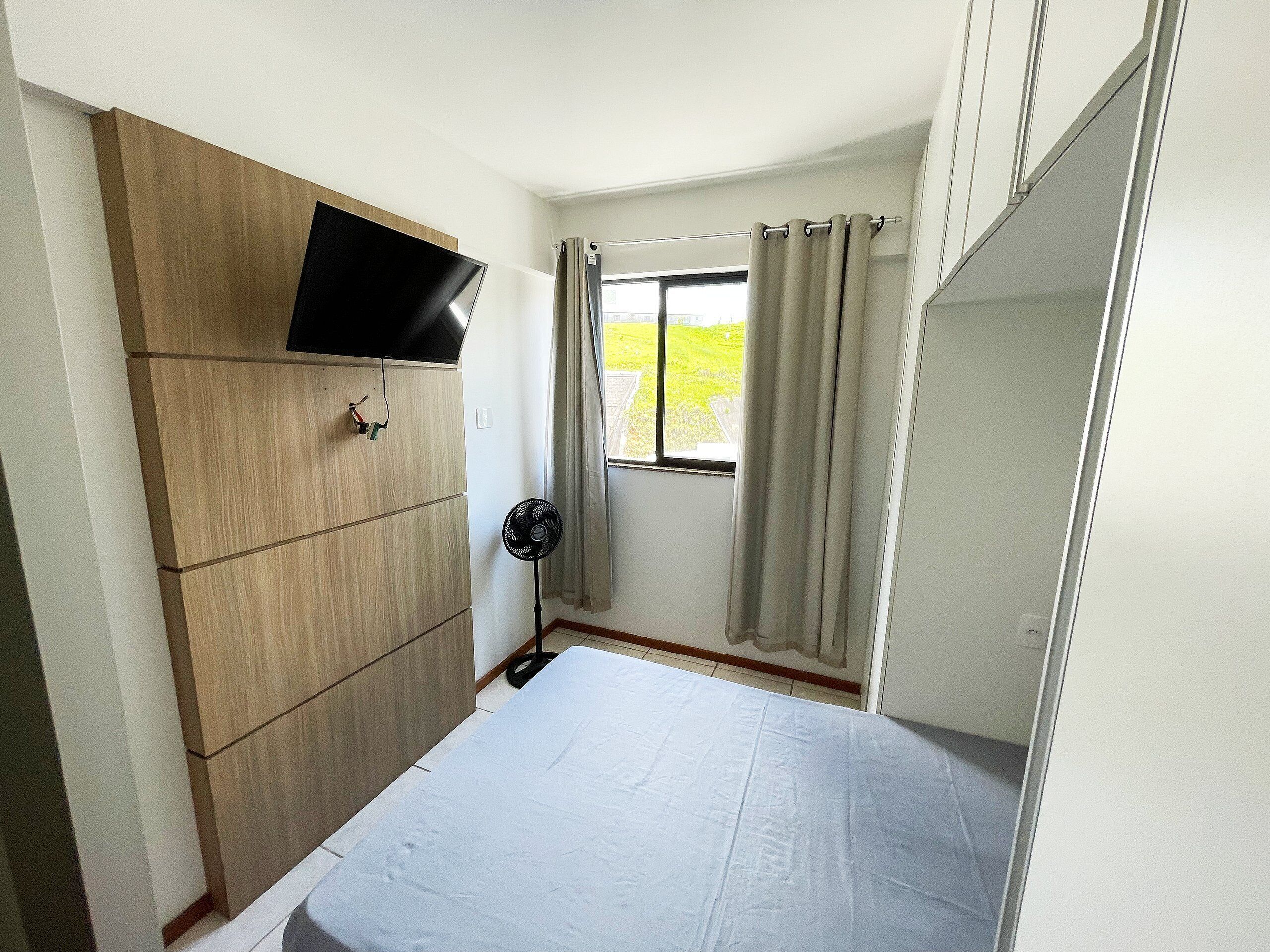 Ein Schlafzimmer mit einem Bett, einem an einem Holzkasten montierten Fernseher, einem Standventilator und einem Fenster mit Vorhängen.
