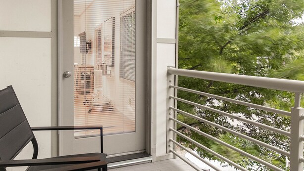 A balcony with a metal railing, a door with blinds, and a view of trees and a building in the background.