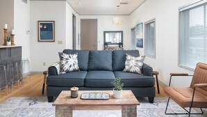 A modern living room with a dark grey sofa, a wooden coffee table, and a potted plant. There is a dining area in the background with a wooden table and chairs.