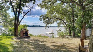 A lakeside outdoor area with a brick fire pit, picnic table, and wooden deck.