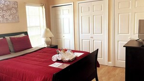 A neatly made bed with a red bedspread, a bedside lamp, and a small table with a folded towel and a basket. - Room in House - Modern Executive Master Bedroom (Alexandria)