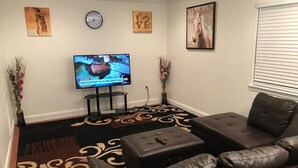 A living room with a leather sofa, a flat-screen TV mounted on the wall, a clock, and two vases with flowers. - Room in House - Modern Executive Master Bedroom (Alexandria)