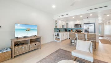 A modern living room with a television, wicker furniture, and a kitchen area with white cabinets and a tiled backsplash.