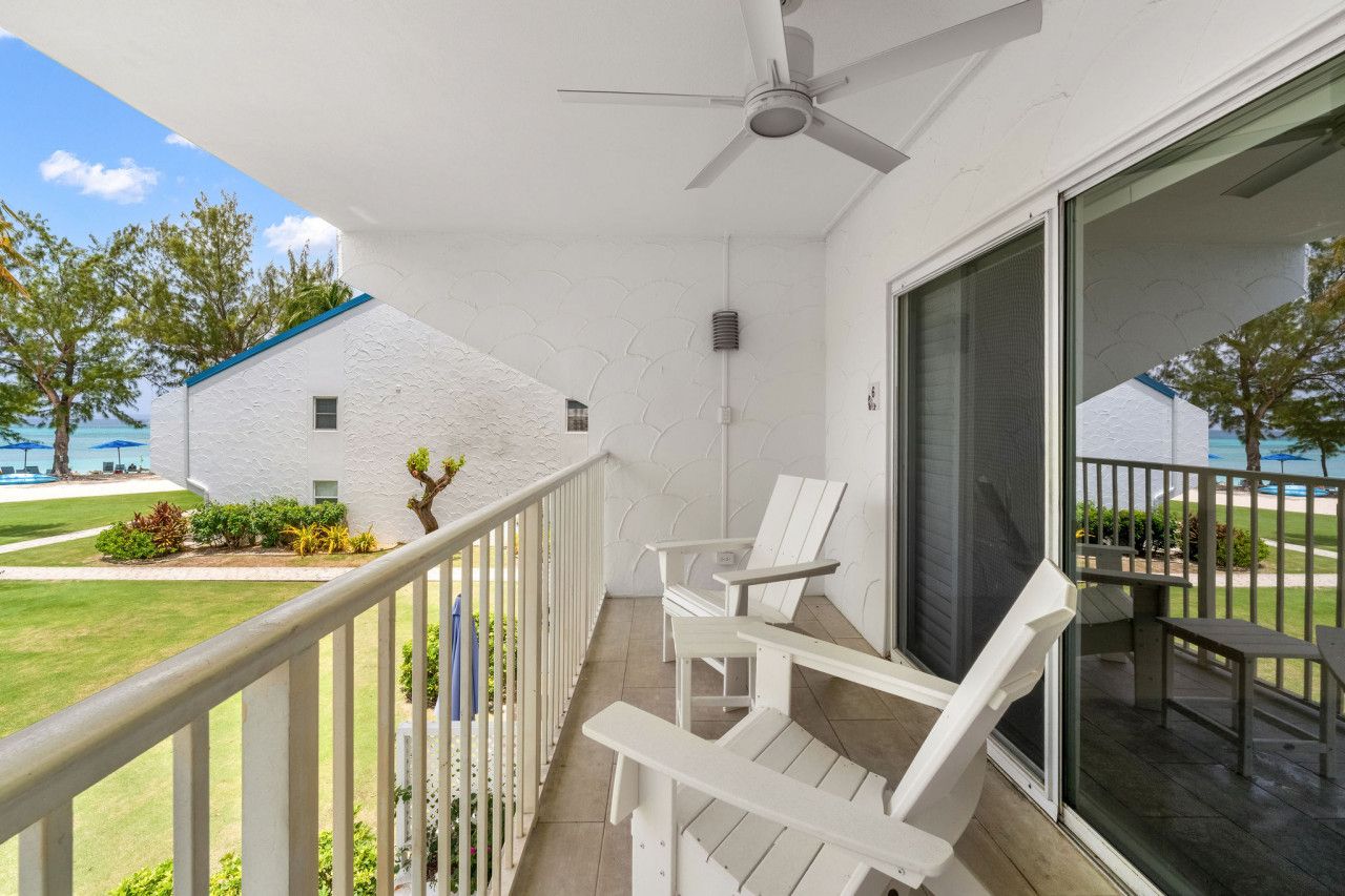 A balcony with white chairs and a ceiling fan, overlooking a lawn and a building with a blue roof.