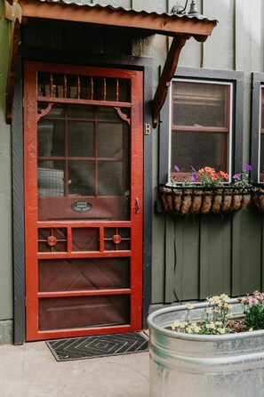 Una puerta roja con un panel de vidrio, un felpudo de metal y una ventana con una maceta colgante de flores.