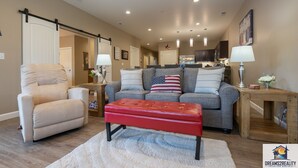 A spacious living room with a gray sofa, red ottoman, and beige armchair. A wooden coffee table and a white rug are in the center. The room has a large sliding door leading to a kitchen and dining area. - Foothills G-104 (Branson)