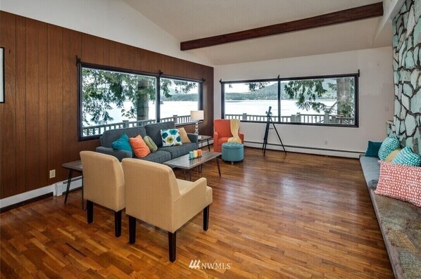 A spacious living room with a wooden floor, a grey sofa with colorful cushions, a beige armchair, and a blue ottoman. Large windows offer a view of a lake and trees.