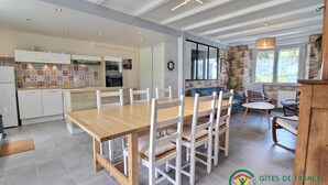 A modern kitchen with white cabinets and a tiled backsplash, a dining area with a wooden table and white chairs, and a stone wall with a window overlooking a garden. - L'avocette - Damgan - Gîte (DAMGAN)
