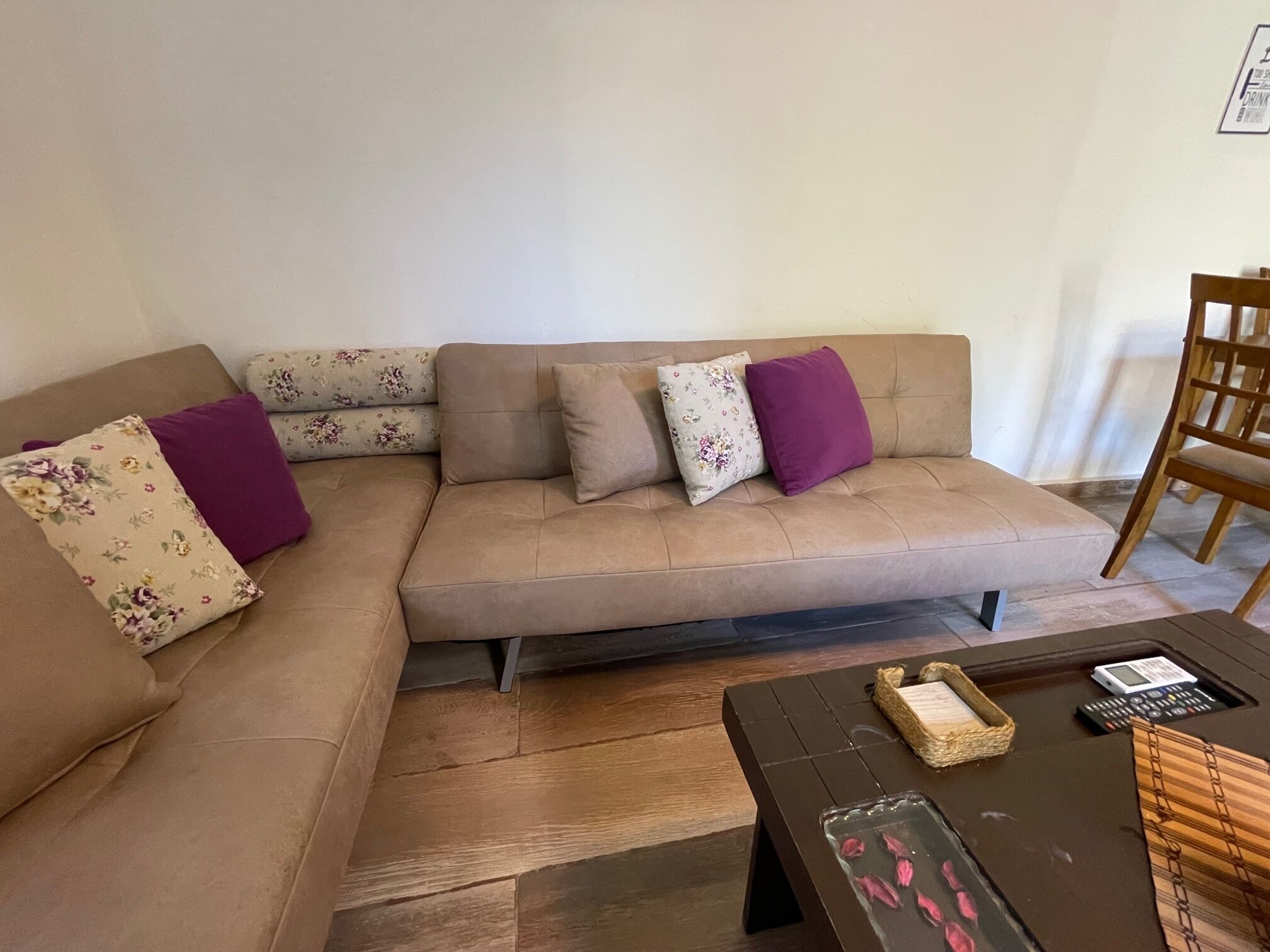 Un sofá seccional color beige con cojines florales y morados sólidos, una mesa de centro de madera oscura con una cesta tejida y un pequeño dispositivo electrónico, y una silla de madera.