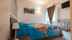 Un dormitorio con una cama, una mesita de noche con una lámpara, un vaso de jugo de naranja, un cuadro en la pared, una ventana con cortinas y un televisor montado en la pared.