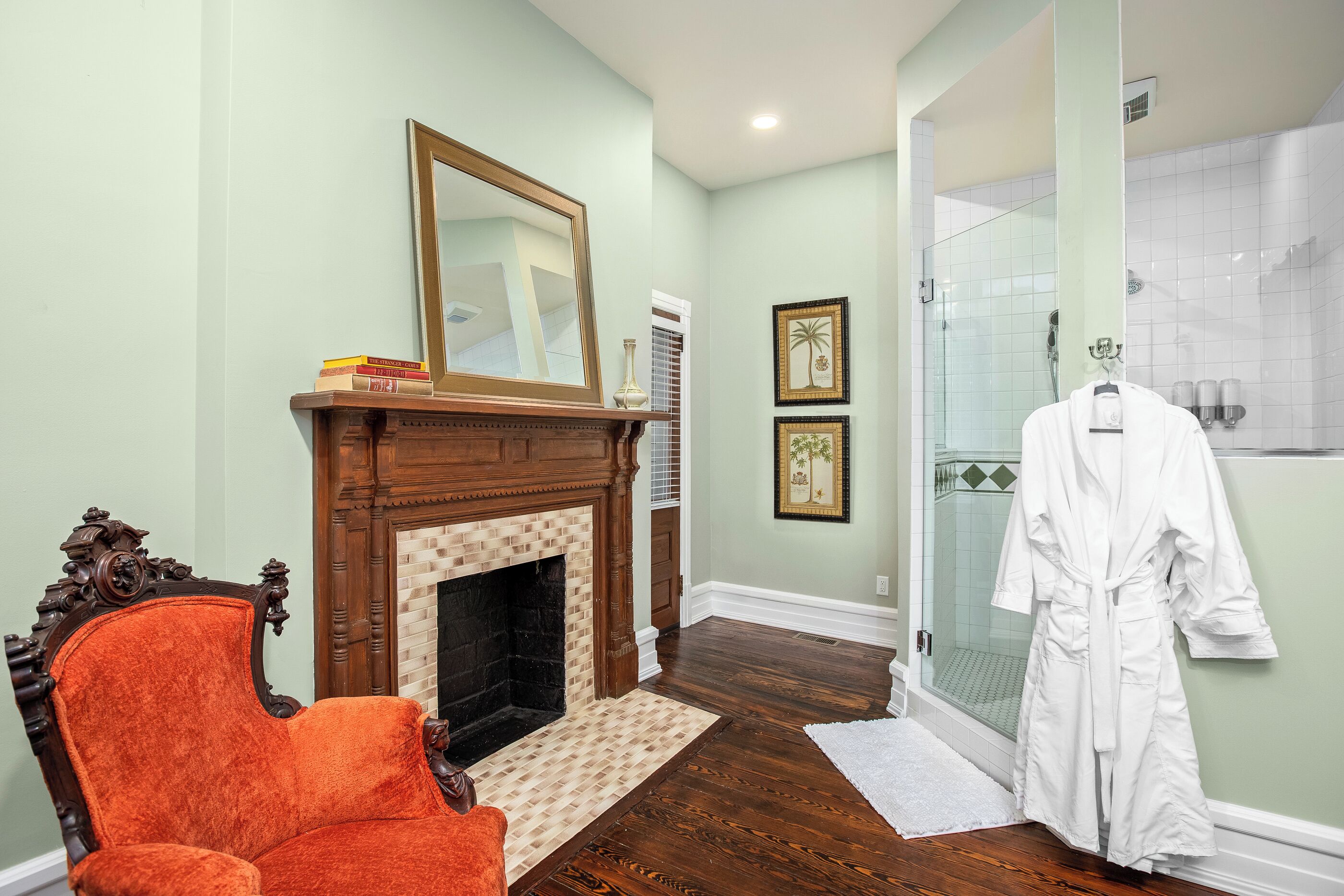 A bathroom with a glass shower, a white robe hanging, and a fireplace with books on a side table.