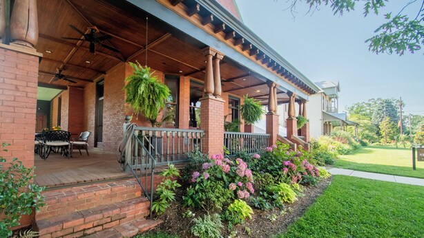A covered porch with brick columns, outdoor seating, and a well-maintained garden with pink flowers. - Summit Street Inns (Winston-Salem)