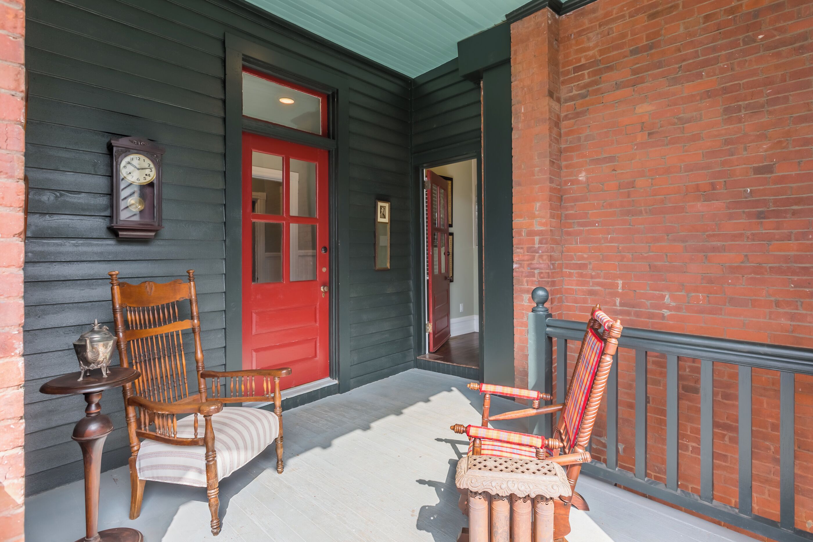 A porch with a red door, a wooden chair, a small table with a lamp, and a brick wall.