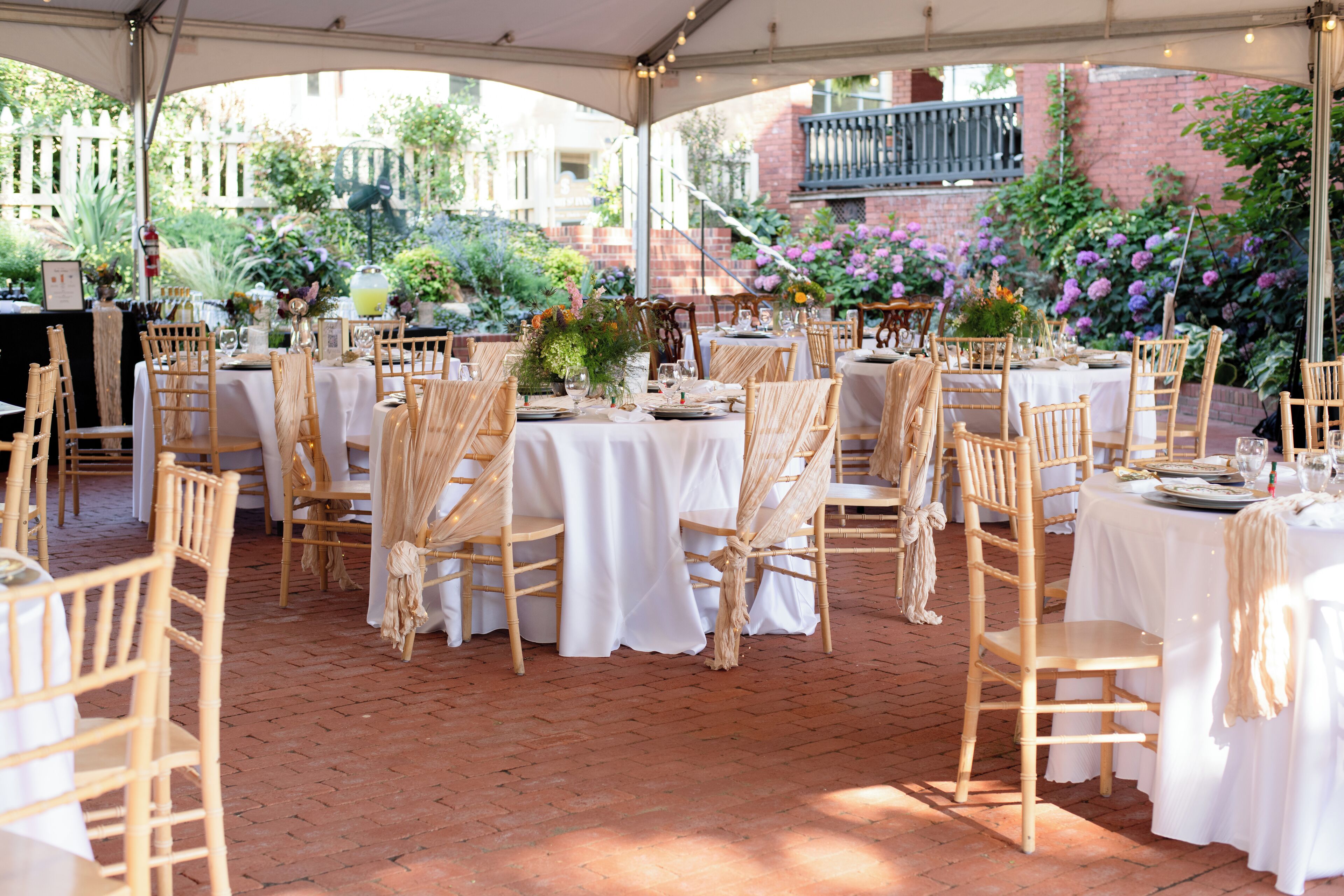 An outdoor dining area with tables set for a meal, surrounded by brick flooring and wooden chairs.