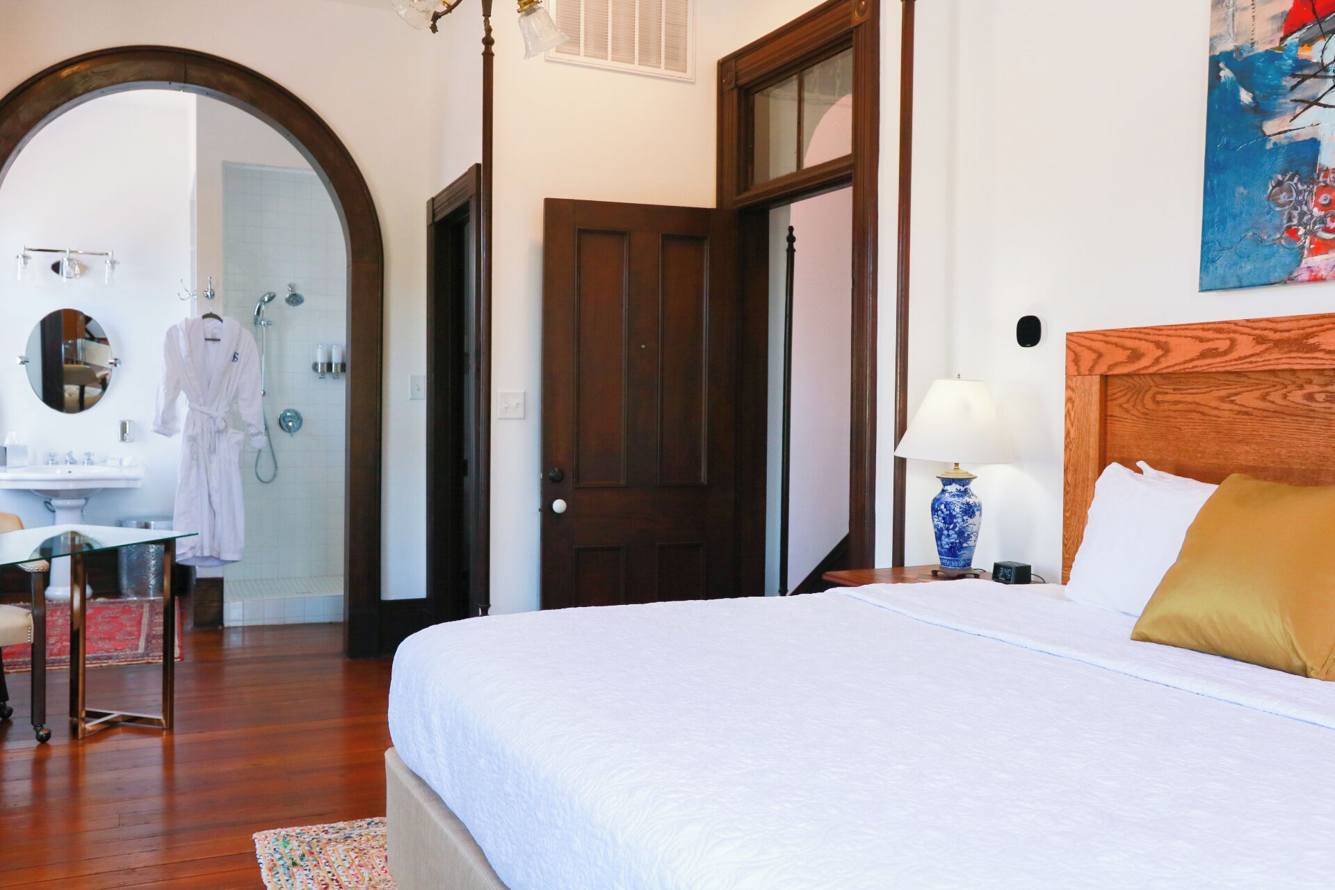 Una camera d'albergo con un letto grande, una testiera in legno, un comodino con una lampada, uno specchio, un bagno con doccia e un accappatoio appeso a un appendiabiti.