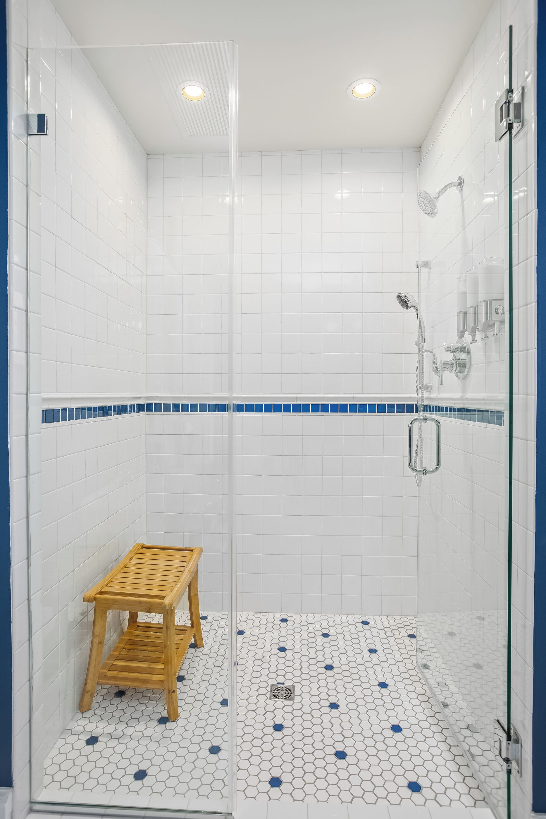 Ein modernes Duschbad mit weißen Fliesen, einem blauen Friesbereich und einem hölzernen Hocker.