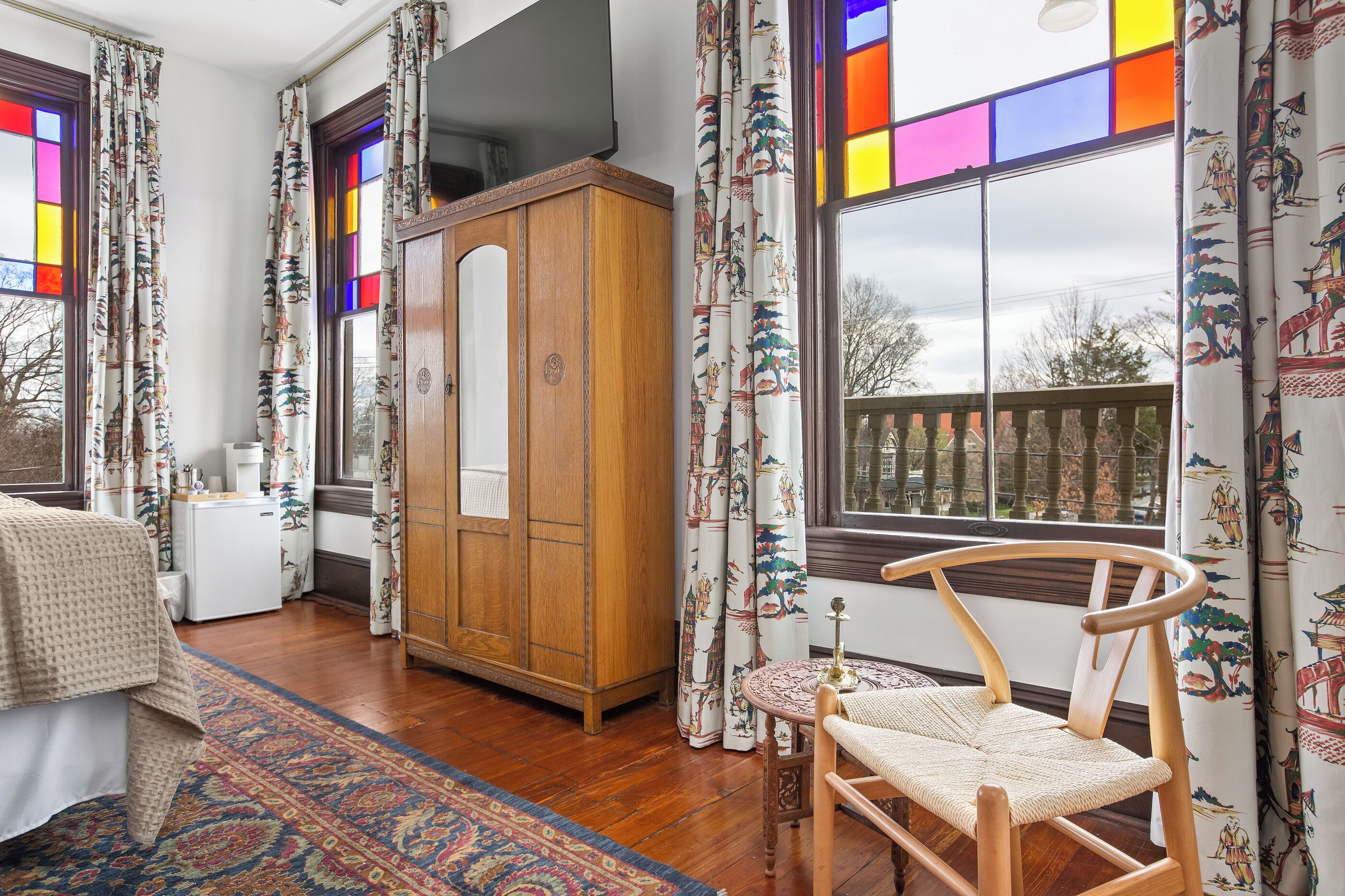 A room with a wooden wardrobe, a round wooden chair, a patterned rug, and a view through a window with colorful stained glass.