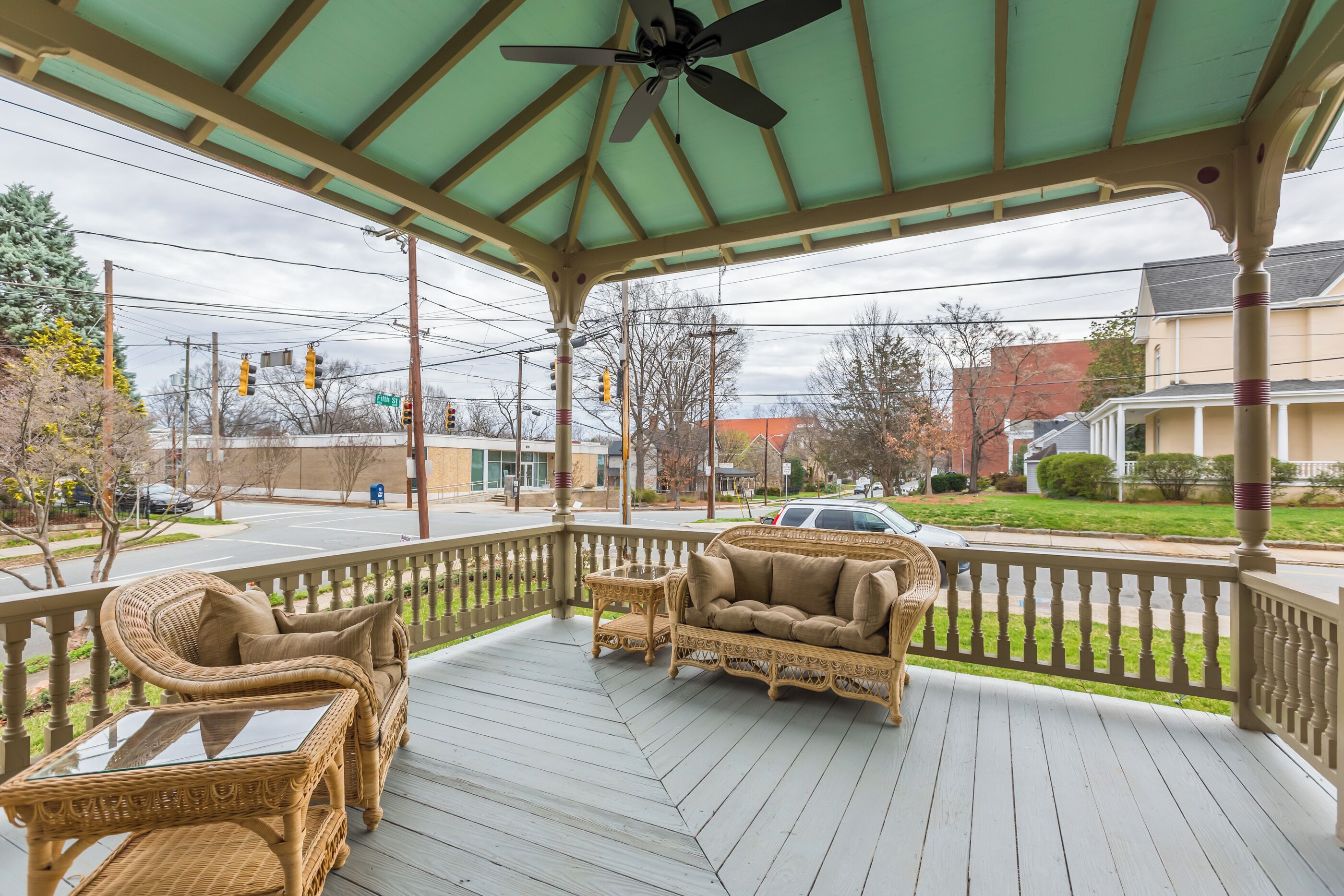 A covered porch with wicker furniture and a glass table, overlooking a street with traffic lights and buildings.