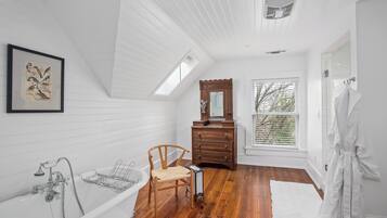 A spacious bathroom with a freestanding bathtub, a wooden chair, a dresser with a mirror, and a large window providing natural light.