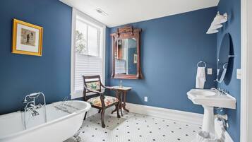 A bathroom with a clawfoot bathtub, a wooden chair, a mirror, and a sink.
