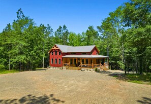 A red cabin with a porch and a gravel driveway, surrounded by trees.