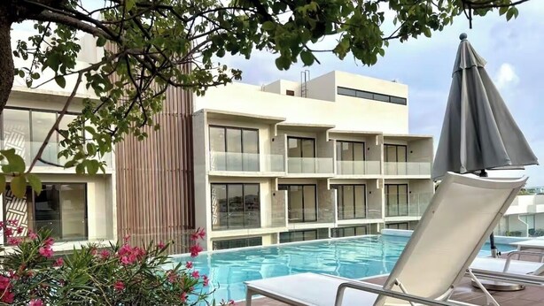 A poolside area with a lounge chair and umbrella, a building with large windows, and a tree with pink flowers. - Departamento con Jacuzzi en Playa del Carmen  (Playa del Carmen)