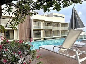 A poolside area with a lounge chair and umbrella, a building with large windows, and a tree with pink flowers.