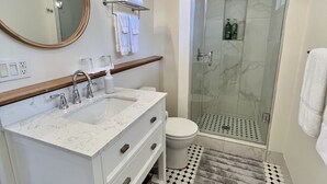 A bathroom with a white vanity, a round mirror, a glass shower enclosure, and a patterned rug. - Mango Sunset Bed and Breakfast (Kailua-Kona)