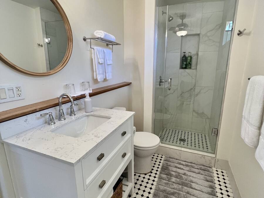 Une salle de bain avec un meuble-lavabo blanc, un miroir rond, une cabine de douche en verre et un tapis à motifs.