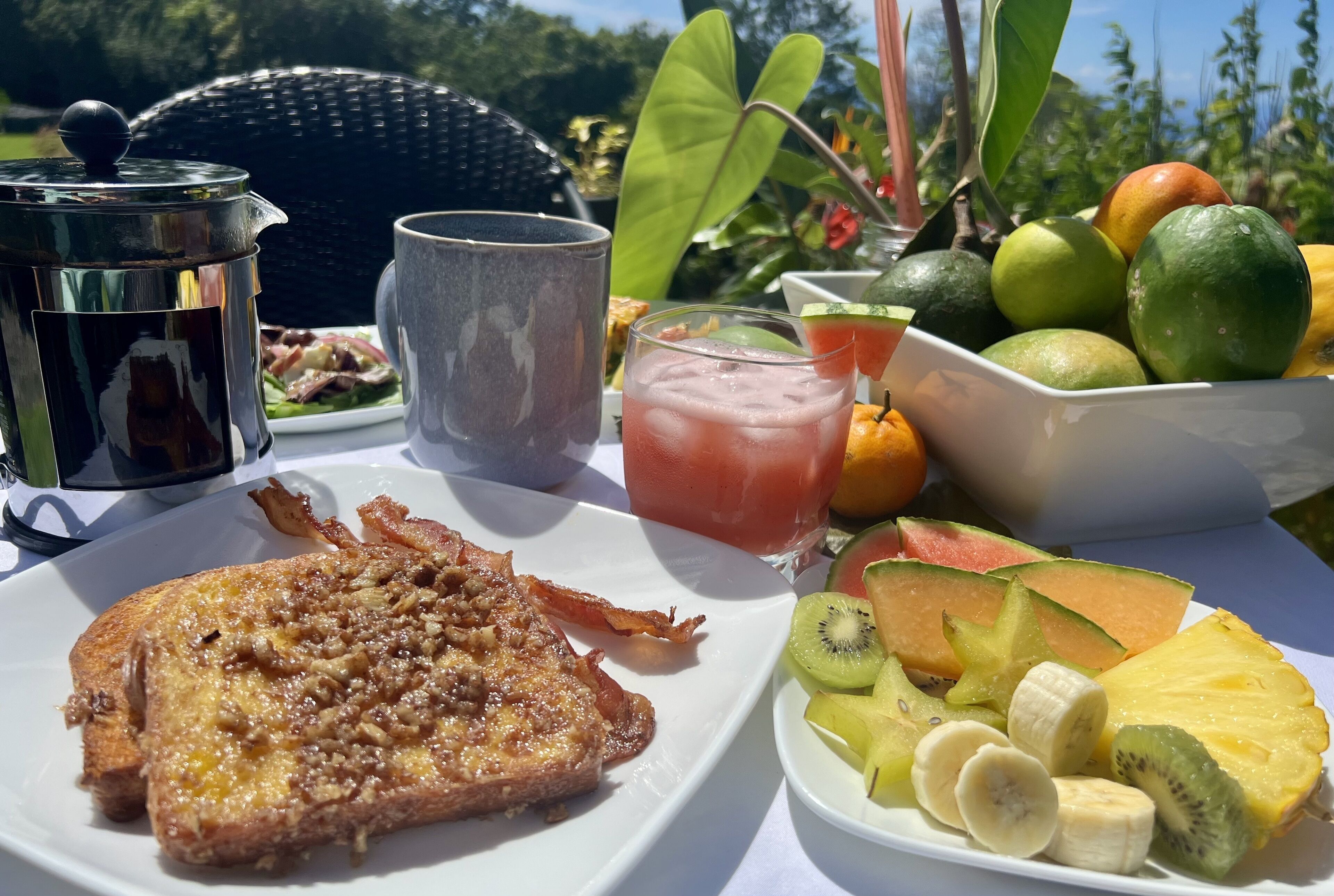 Une table dressée pour le petit-déjeuner, avec des toasts, du bacon, des fruits et une boisson, le tout installé à l’extérieur.