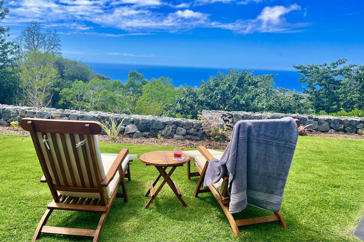 A wooden deck with two chairs and a table, overlooking a grassy area and a stone wall.