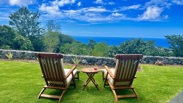 Two wooden chairs with cushions, a small round table, and a stone wall. - Mango Sunset Bed and Breakfast (Kailua-Kona)