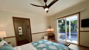 A hotel room with a bed, a ceiling fan, a TV, and a view of the garden through the sliding glass door. - Mango Sunset Bed and Breakfast (Kailua-Kona)