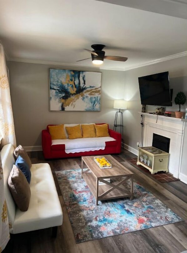 A living room with a red sofa, a white armchair, a coffee table, a colorful rug, a fireplace, a television, and a ceiling fan.
